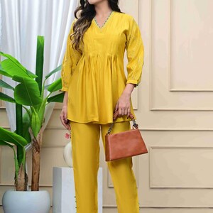 May include: Mustard yellow two-piece outfit featuring a V-neck top with gathered detailing and matching pants. The top has three-quarter sleeves with embroidered accents. A brown handbag with a woven handle and a pair of clear heels complete the ensemble.