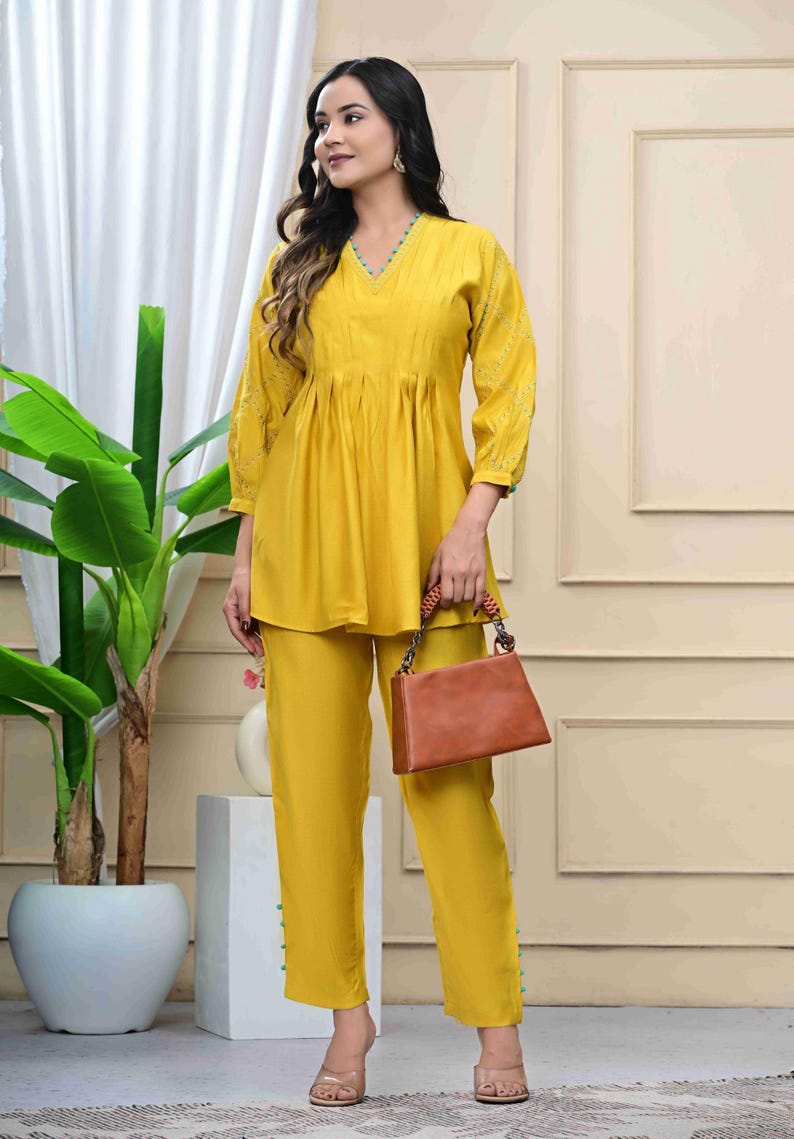 May include: Mustard yellow two-piece outfit featuring a V-neck top with gathered detailing and matching pants. The top has three-quarter sleeves with embroidered accents. A brown handbag with a woven handle and a pair of clear heels complete the ensemble.