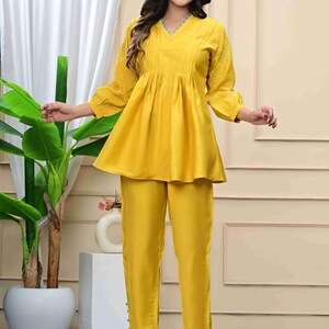 May include: A bright yellow two-piece outfit. The top has a V-neck, gathered waist, and three-quarter sleeves with embroidered details. The matching pants are straight-legged. The outfit is made of a smooth, flowing fabric.