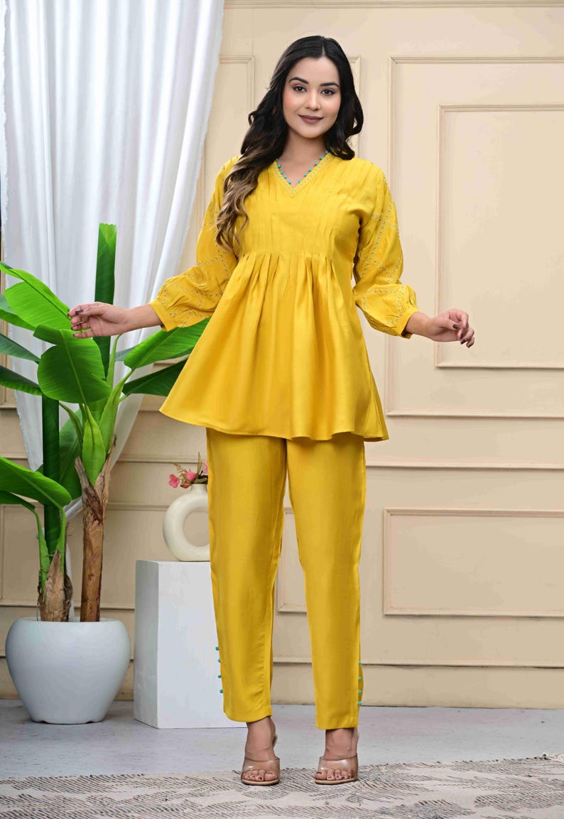 May include: A bright yellow two-piece outfit. The top has a V-neck, gathered waist, and three-quarter sleeves with embroidered details. The matching pants are straight-legged. The outfit is made of a smooth, flowing fabric.