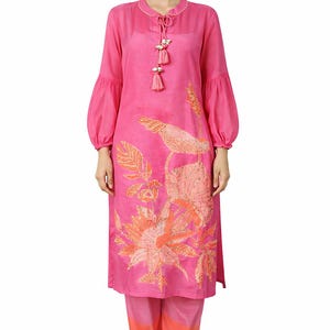 Puede incluir: Un conjunto de dos piezas rosa vibrante, compuesto por una túnica de manga larga y pantalones a juego. La túnica presenta un diseño floral en tonos naranja y melocotón. Los pantalones hacen una transición de rosa a naranja. El conjunto incluye borlas decorativas.