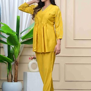 May include: A mustard yellow two-piece outfit. The top has a V-neck, long sleeves with embroidered details, and a gathered waist. The matching pants are straight-legged. The outfit is worn with clear heels.