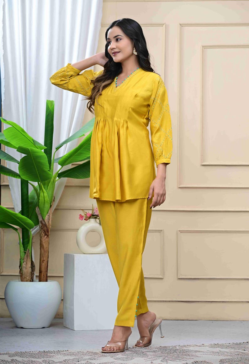 May include: A mustard yellow two-piece outfit. The top has a V-neck, long sleeves with embroidered details, and a gathered waist. The matching pants are straight-legged. The outfit is worn with clear heels.