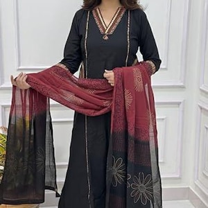 Puede incluir: Conjunto de salwar kameez negro con un dupatta burdeos y negro. La túnica de manga larga presenta adornos dorados en el escote y en la parte delantera. El dupatta tiene un estampado floral y detalles dorados. El conjunto se usa con pantalones palazzo negros.