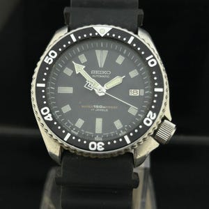 May include: A black Seiko automatic wristwatch with a black dial, luminous hands, and a date display at the 3 o'clock position. The watch has a black bezel with white markings and a black rubber strap. The watch is water resistant to 150m.