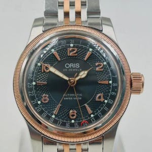 Może przedstawiać: Srebrny i różowo-złoty zegarek Oris z ciemnozieloną tarczą. Zegarek posiada bransoletę ze stali nierdzewnej, różowo-złotą ramkę i napis "ORIS 25 JEWELS AUTOMATIC SWISS MADE".