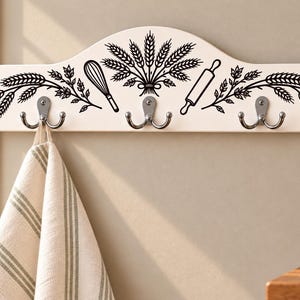 May include: A white wall-mounted rack with three silver double hooks. The rack features a black graphic print of wheat stalks, a wire whisk, and a rolling pin. A beige and white striped kitchen towel hangs from the leftmost hook on the rack.