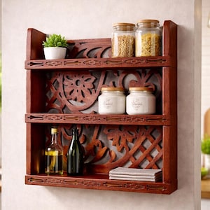 Peut inclure: Étagère murale en bois brun foncé à trois niveaux, avec des motifs floraux et géométriques sculptés. Les étagères contiennent des bocaux, des bouteilles, une petite plante et un tissu plié. L'étagère est fixée à un mur clair.