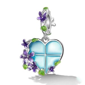 May include: A silver heart-shaped charm pendant with a light blue centre divided into four sections. The charm is decorated with purple flowers and green leaves. The pendant is designed to be attached to a bracelet or necklace.