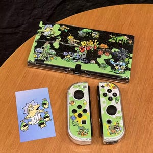 Op de afbeelding: Een Nintendo Switch-console en Joy-Con-controllers met een aangepast ontwerp. De consolebehuizing is zwart en groen met cartoonafbeeldingen en de tekst "Work's Over!". De Joy-Cons zijn groen en wit met bijpassende illustraties. Er is ook een kleine kaart met een cartoon aanwezig.