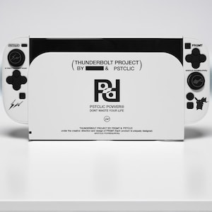 Pode incluir: Uma consola de jogos branca e preta com o texto "(THUNDERBOLT PROJECT) BY & PSTCLIC" e "PSTCLIC POWER DON'T WASTE YOUR LIFE". A consola tem botões pretos e um design de raio. O design é de FRGMT & PSTCLIC.