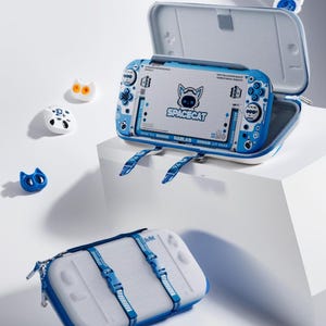 May include: A white and blue gaming console case with a Spacecat design and matching accessories. The open case displays the console with a cat logo. Several cat-shaped accessories are placed around the case.