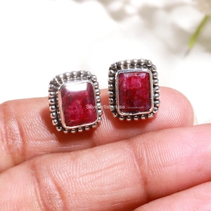 May include: A pair of rectangular stud earrings featuring deep red, faceted gemstones set in silver frames. Each setting is bordered by a decorative beaded metal edge. The text Silverartistusa is visible on the surface of the left gemstone.