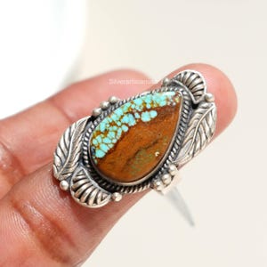 May include: A sterling silver ring featuring a pear-shaped turquoise stone with brown matrix. The setting is decorated with embossed leaf motifs and small silver spheres along the edges. The text Silverartisianusa is visible on the ring surface.