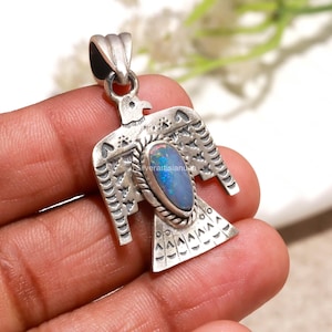 May include: A silver pendant shaped like a bird with outstretched wings, featuring a central teardrop-shaped iridescent blue opal stone. The metal surface is oxidized with stamped geometric patterns and a rope-textured border.