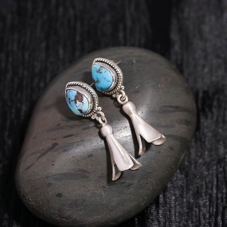 Peut inclure: Une paire de boucles d'oreilles en argent avec des pierres turquoise. Les boucles d'oreilles pr&eacute;sentent un design d&eacute;taill&eacute; autour des pierres turquoise et un charm en forme de cloche. Les boucles d'oreilles sont pr&eacute;sent&eacute;es sur une pierre grise fonc&eacute;e.