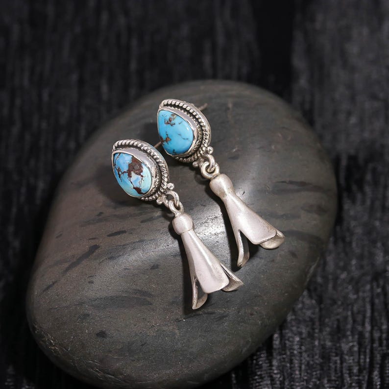Peut inclure: Boucles d'oreilles en argent avec des pierres turquoise. Les boucles d'oreilles pr&eacute;sentent un design d&eacute;taill&eacute; autour des pierres turquoise et un charm en forme de cloche. Elles sont pr&eacute;sent&eacute;es sur une pierre grise fonc&eacute;e.
