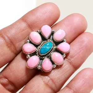 May include: A silver ring featuring a central oval turquoise stone surrounded by a twisted rope border and eight teardrop-shaped pink stones arranged in a floral pattern. The metal setting has a polished finish with small silver bead accents.