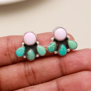 May include: A pair of stud earrings featuring a round, light pink stone at the top, accented by three teardrop-shaped turquoise stones in a silver setting. The text silverartisanusa is visible on the metal surface between the earrings.