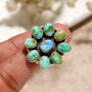 May include: A silver ring featuring a central oval turquoise stone surrounded by a twisted metal border and eight smaller oval turquoise stones in varying shades of blue and green. The text silverartisanusa is visible on the center stone.
