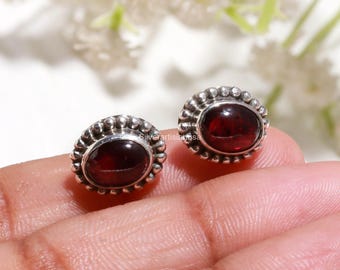 Op de afbeelding: Een paar ovale oorknopjes met dieprode granaat edelstenen gezet in gepolijst zilver. Elke steen is omringd door een decoratieve kralenrand van zilver, wat een textuurrijk kader vormt rond de gladde, donkerrode cabochon-middelpunten.