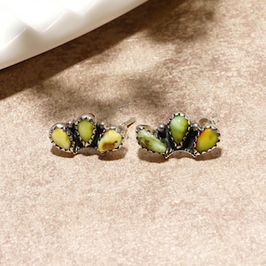 May include: A pair of sterling silver stud earrings featuring three teardrop-shaped, variegated light green and yellow stones set in a serrated metal bezel. The earrings have a dark, oxidized finish around the stones and include post backs.