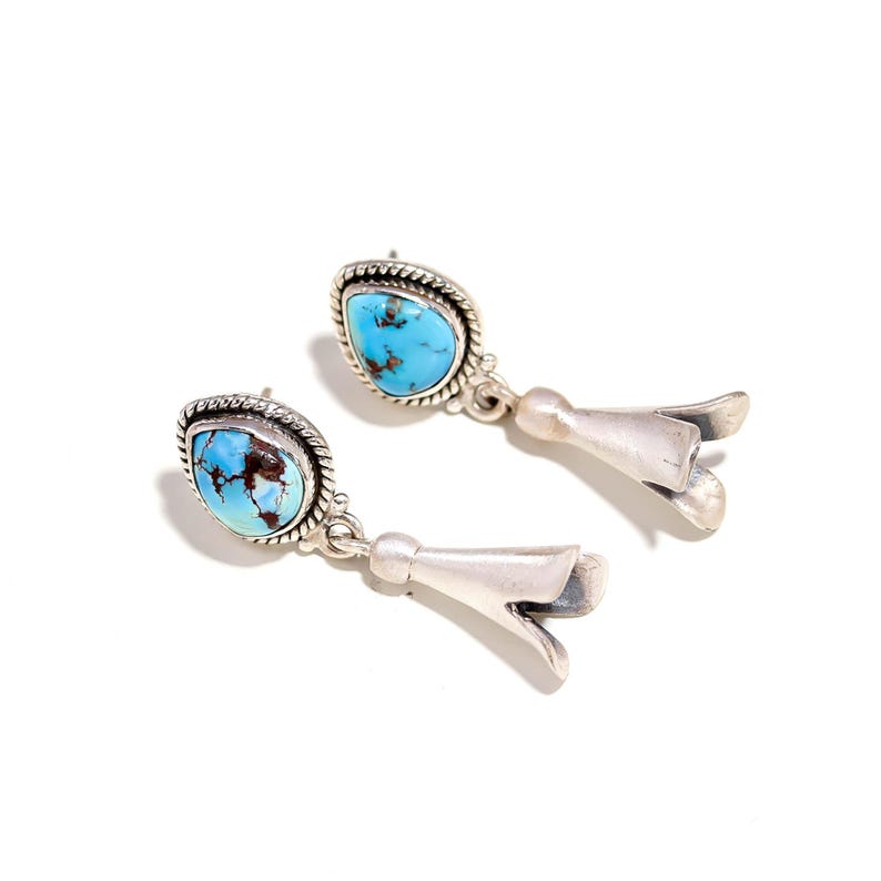 Peut inclure: Une paire de boucles d'oreilles en argent avec des pierres turquoise. Chaque boucle d'oreille pr&eacute;sente une pierre turquoise en forme de goutte d'eau sertie dans une lunette en argent, avec une fleur d&eacute;corative en argent en dessous.