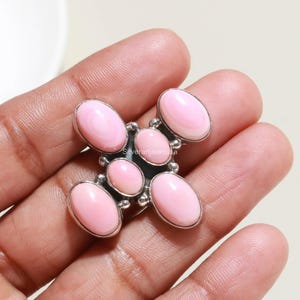 May include: A silver ring featuring five oval-shaped, light pink cabochon stones arranged in a cross pattern. The stones are set in polished silver bezels with small silver bead accents between each stone. The text Silverartisianusa is visible.