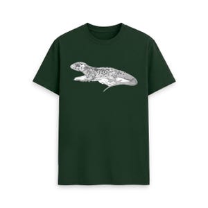 May include: A dark forest green short-sleeved t-shirt featuring a detailed black and white line art illustration of a lizard resting on a rock centred on the chest. The graphic depicts the reptile in profile with textured scales and a long tail.