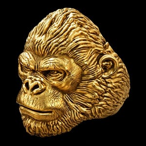 May include: A gold-colored ring featuring a detailed, three-dimensional sculpture of a gorilla head. The design captures the primate's facial features, including the brow, nose, and mouth, with textured fur patterns covering the entire surface.