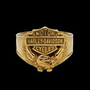Può includere: Un anello color oro con il logo Harley-Davidson al centro. Il design include una forma a scudo con la scritta MOTOR HARLEY-DAVIDSON CYCLES e un dettagliato emblema a forma di aquila posizionato proprio sotto lo scudo, integrato nella fascia.