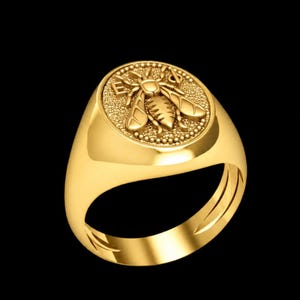 Può includere: Un anello con sigillo d'oro con un dettagliato emblema a forma di ape. L'anello ha una finitura lucida e una faccia arrotondata con il disegno dell'ape al centro. La parola "EYES" è incisa intorno all'ape. L'anello è su sfondo nero.
