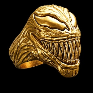 Könnte beinhalten: Ein goldfarbener Ring mit einem skulptierten, detaillierten Kopf, großen, mandelförmigen Augen und einem breiten, grinsenden Mund voller spitzer Zähne. Die Oberfläche des Rings hat ein strukturiertes, organisches Aussehen mit tiefen Rillen.