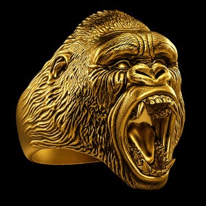 May include: A gold-toned ring featuring a highly detailed, three-dimensional sculpture of a gorilla head. The gorilla is depicted with an open mouth, bared teeth, and textured fur detailing across the entire surface of the metal band.