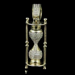 May include: A gold-colored hourglass pendant encrusted with small, sparkling stones. The hourglass is held within a decorative gold frame with a loop at the top for a chain. The pendant is set against a black background.