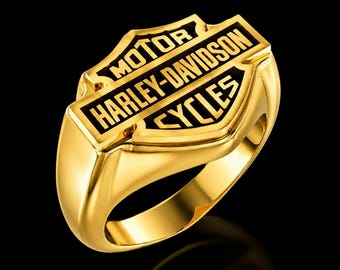 May include: A polished gold-tone ring featuring the Harley-Davidson Motor Cycles logo on the face. The emblem is raised with black enamel detailing against the metallic gold surface, showcasing the iconic shield and bar design clearly.