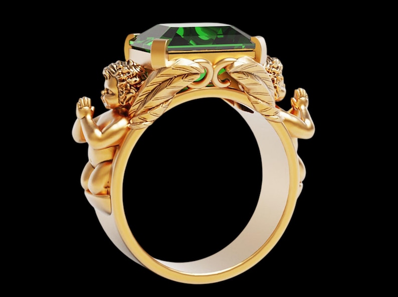 Peut inclure: Bague en or orn&eacute;e d'une grande pierre verte taill&eacute;e en &eacute;meraude. Deux ch&eacute;rubins encadrent la gemme, avec des d&eacute;tails de plumes. La bague pr&eacute;sente un design bicolore, avec un ext&eacute;rieur dor&eacute; et un int&eacute;rieur argent&eacute;.