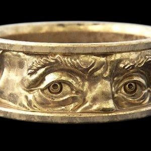 Könnte beinhalten: Ein goldfarbener Ring mit einem detaillierten Relief eines Gesichts, das zwei Augen und einen Teil der Stirn zeigt. Der Ring hat ein Banddesign mit strukturierter Oberfläche vor schwarzem Hintergrund.