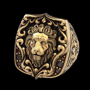 Puede incluir: Un anillo dorado con una detallada cabeza de león con una melena fluida, sobre un fondo negro. El anillo tiene adornos de volutas y pequeños acentos rojos, creando un diseño regio y detallado.