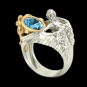 May include: A two-tone ring featuring a central oval-cut light blue gemstone set in a gold-coloured frame. The band is crafted in a silver-coloured metal and depicts a sculpted figure with detailed wings that wrap around the sides of the ring.