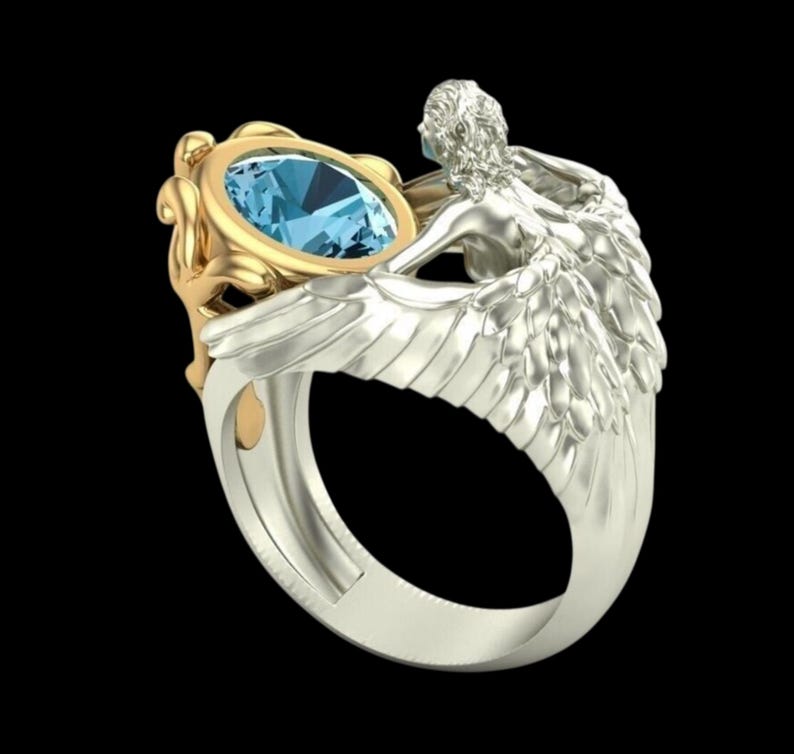 May include: A two-tone ring featuring a central oval-cut light blue gemstone set in a gold-coloured frame. The band is crafted in a silver-coloured metal and depicts a sculpted figure with detailed wings that wrap around the sides of the ring.