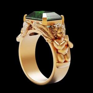 Peut inclure: Une bague en or orn&eacute;e d'une grande pierre verte taill&eacute;e en &eacute;meraude. La monture de la bague est orn&eacute;e de motifs d&eacute;taill&eacute;s de ch&eacute;rubins et de feuilles. La bague est pos&eacute;e sur un fond noir, mettant en valeur ses d&eacute;tails.