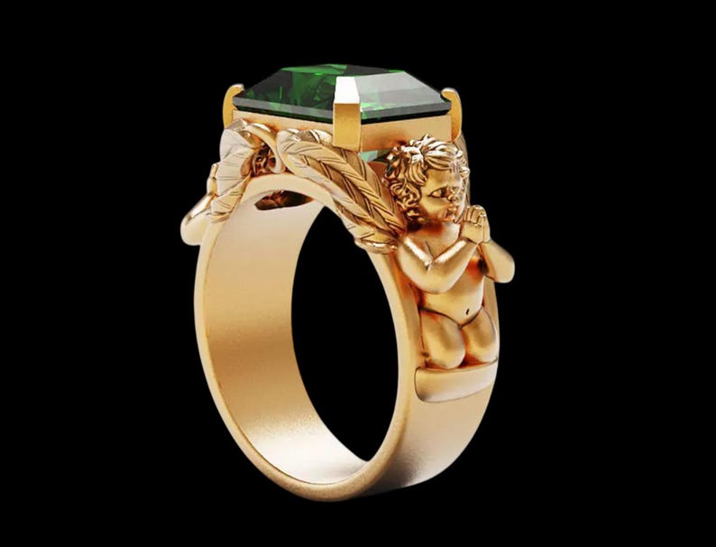 Peut inclure: Une bague en or orn&eacute;e d'une grande pierre verte taill&eacute;e en &eacute;meraude. La monture de la bague est orn&eacute;e de motifs d&eacute;taill&eacute;s de ch&eacute;rubins et de feuilles. La bague est pos&eacute;e sur un fond noir, mettant en valeur ses d&eacute;tails.