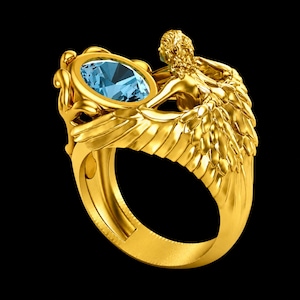 May include: A gold ring featuring a sculpted figure with detailed wings extending along the band. The centrepiece is an oval-cut light blue gemstone held within a decorative setting. The metal has a polished finish and intricate textures.