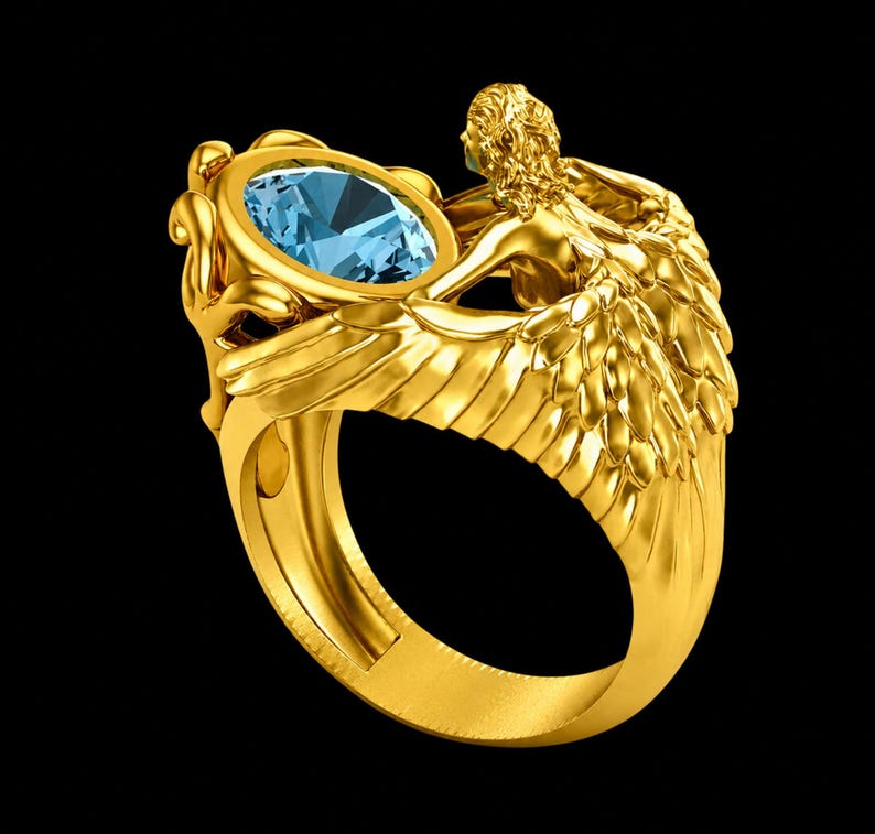 May include: A gold ring featuring a sculpted figure with detailed wings extending along the band. The centrepiece is an oval-cut light blue gemstone held within a decorative setting. The metal has a polished finish and intricate textures.