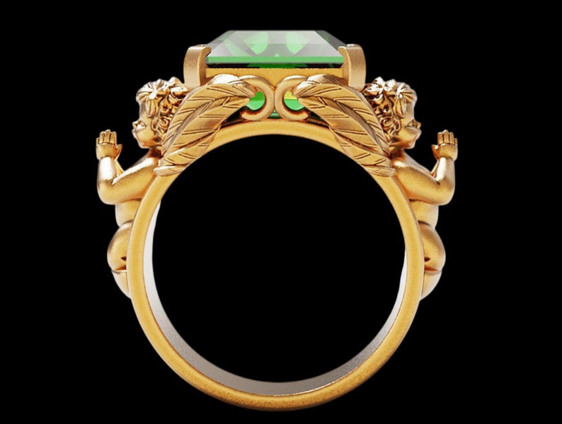 Peut inclure: Une bague en or orn&eacute;e d'une grande pierre verte taill&eacute;e en &eacute;meraude. Deux ch&eacute;rubins aux bras tendus encadrent la gemme, avec des d&eacute;tails de plumes. La bague a une finition polie, sur fond noir.