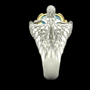 May include: A silver-toned ring featuring a sculpted figure with detailed wings extending along the band. The figure is positioned above a central setting that holds a round, light blue gemstone encased in a polished gold-toned metal frame.