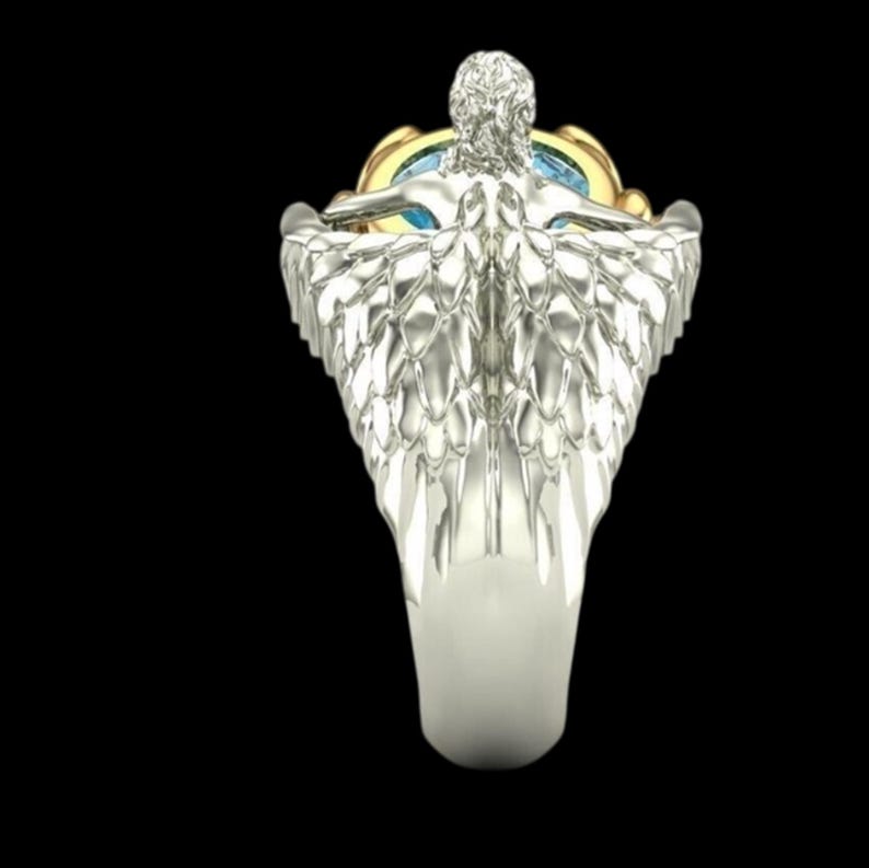 May include: A silver-toned ring featuring a sculpted figure with detailed wings extending along the band. The figure is positioned above a central setting that holds a round, light blue gemstone encased in a polished gold-toned metal frame.