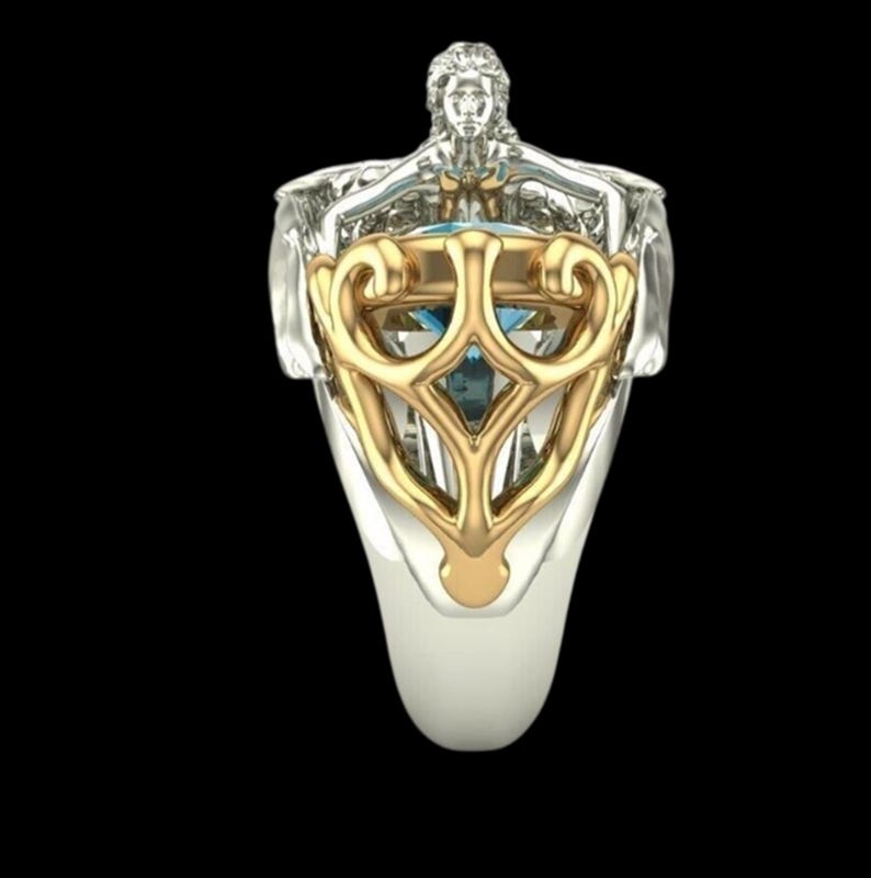 May include: A silver-toned ring featuring a sculpted human figure at the top, with arms extended over a central blue gemstone. The stone is framed by an ornate, gold-toned metal overlay with curved, symmetrical scrollwork patterns.
