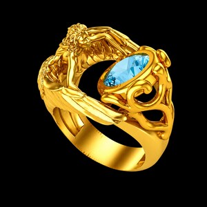 May include: A gold ring featuring a sculpted figure with wings that wrap around the band. The ring is set with a single, oval-cut, light blue gemstone held within a decorative, swirling gold setting that connects to the winged figure's form.